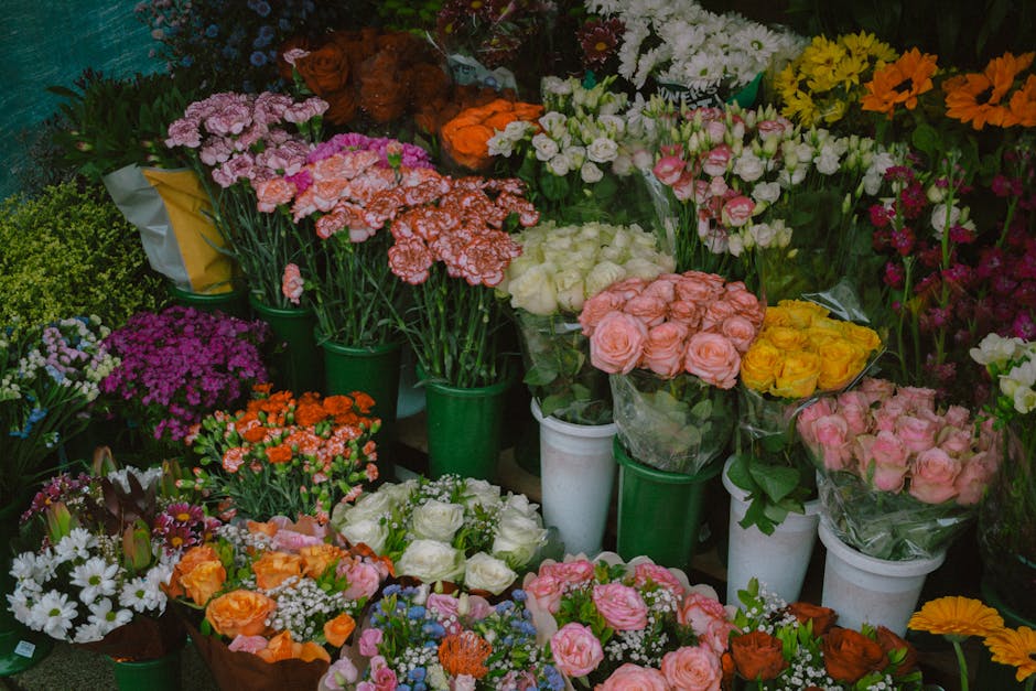 A diverse array of fresh flowers arranged in green, white, and clear wrapping, including pink, yellow, white, orange, and purple blooms such as roses, carnations, and daisies. The flowers are placed i