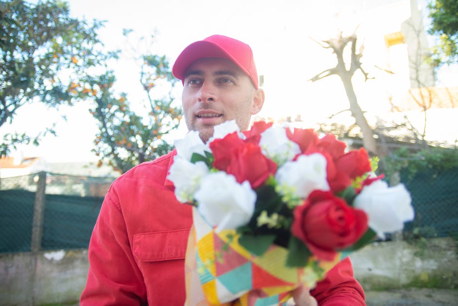 A young man dressed in a red jacket and matching red cap is holding a vibrant bouquet of fresh roses, including red and white blooms with green foliage, wrapped in colorful patterned paper. He appears