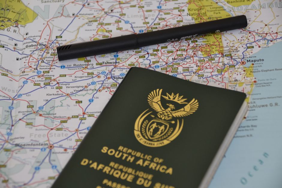 A detailed photograph of a bouquet of fresh flowers resting on a map, accompanied by a black pen and a South African passport. The bouquet features an arrangement of vibrant roses and peonies in shade