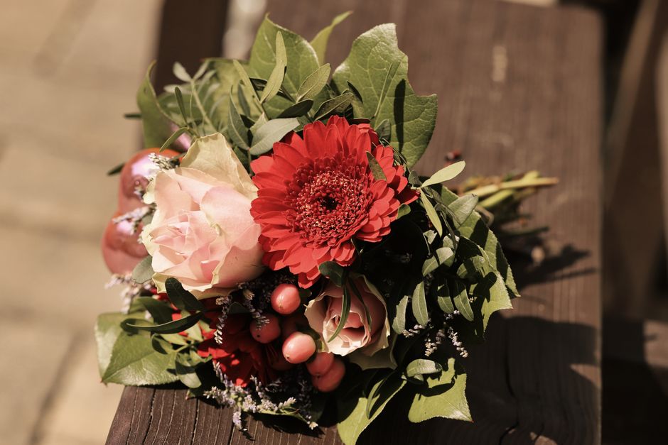 A close-up of a floral bouquet featuring a large coral-red gerbera daisy in the center, surrounded by soft pink roses, small pink hypericum berries, and sprigs of purple veronica, all accented with va